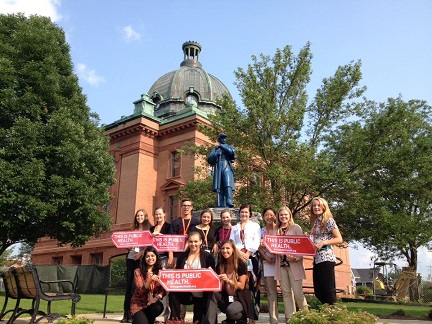 A dozen people stand in front of a building holding red signs that read, THIS IS PUBLIC HEALTH