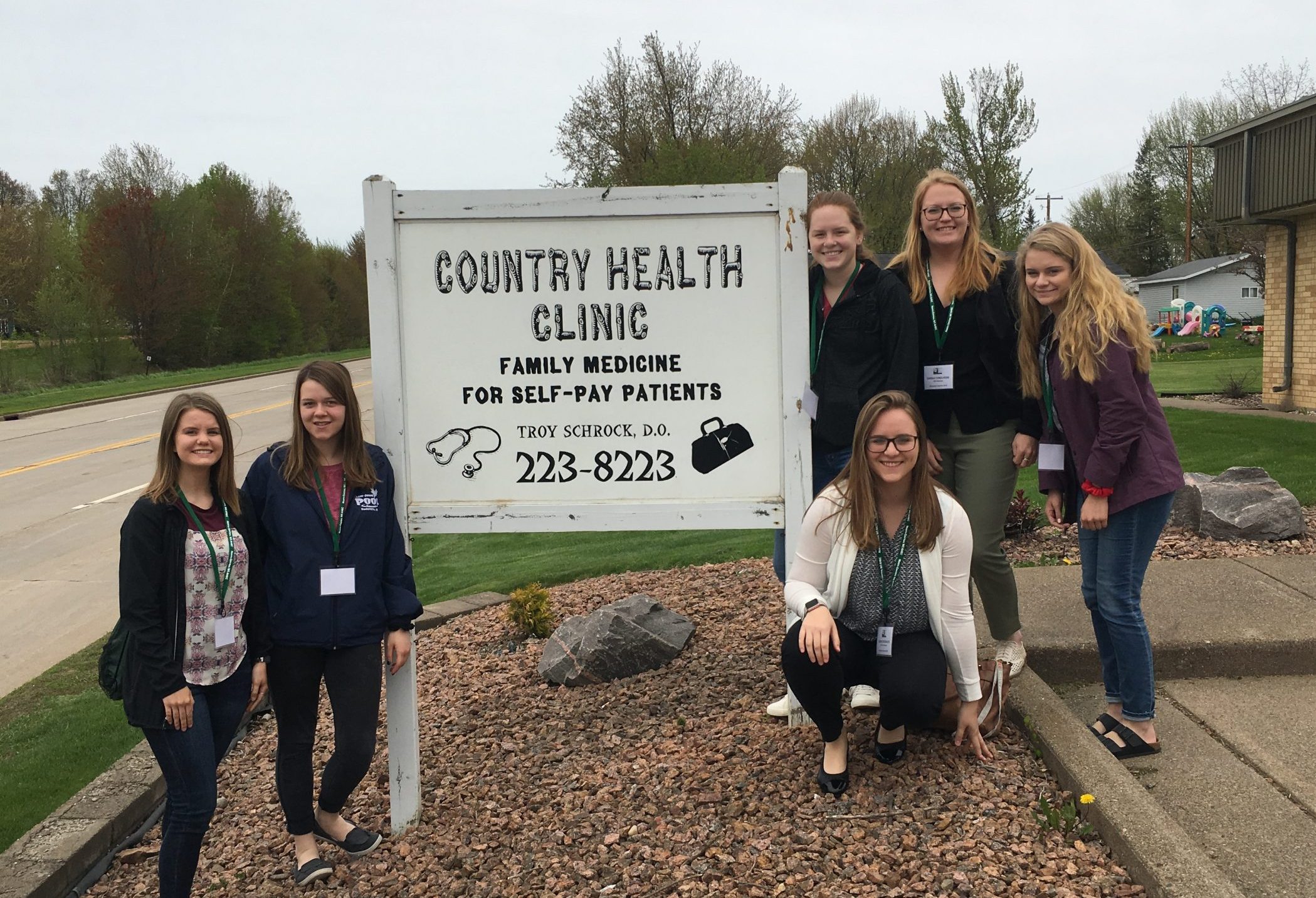 A group of six students with name badges smile and post in front of a sign that reads, "Country Health Clinic. Family medicine for self-pay patients. Troy Schrock D.O."