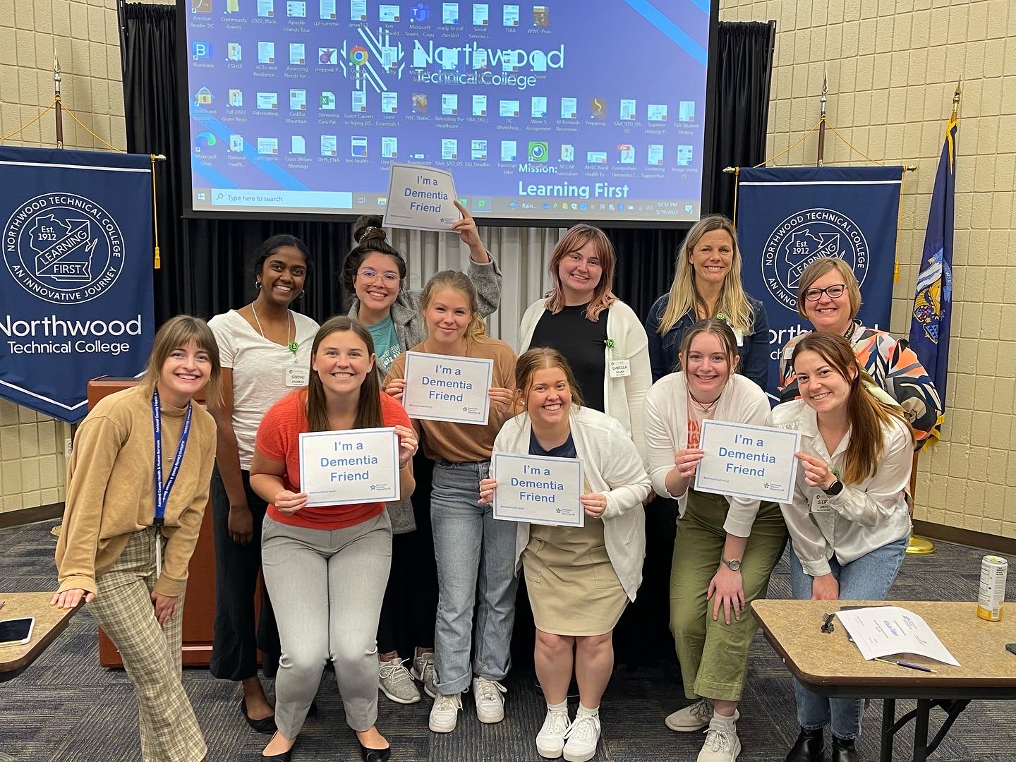 A group of AHEC Scholars hold signs that say "I'm a Dementia Friend" in front of blue flags for Northwood Technical College and a projector screen with a computer desktop