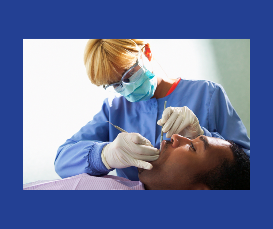 A person with short hair in a blue mask, blue scrubs and white latex gloves performs a dental examination on a man in a reclined position