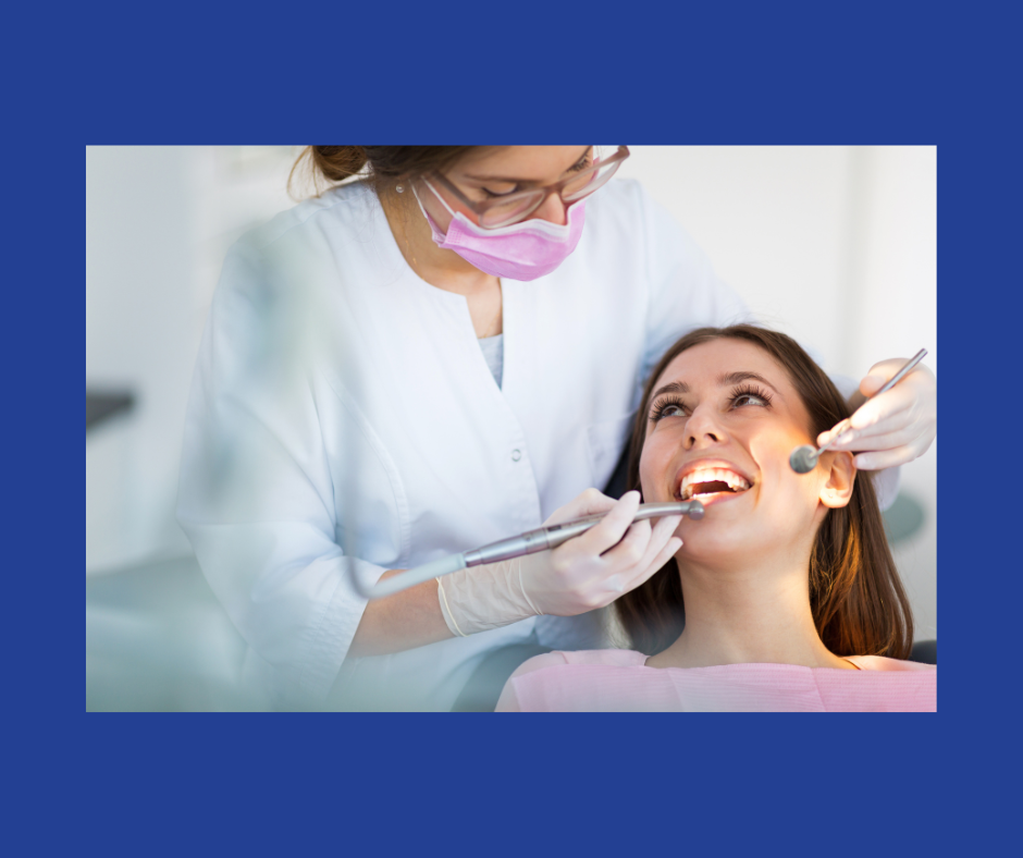 A woman with long dair hair smiles up at a dentist in glasses, a pink face mask and a white coat who shines a light in the mouth of the patient