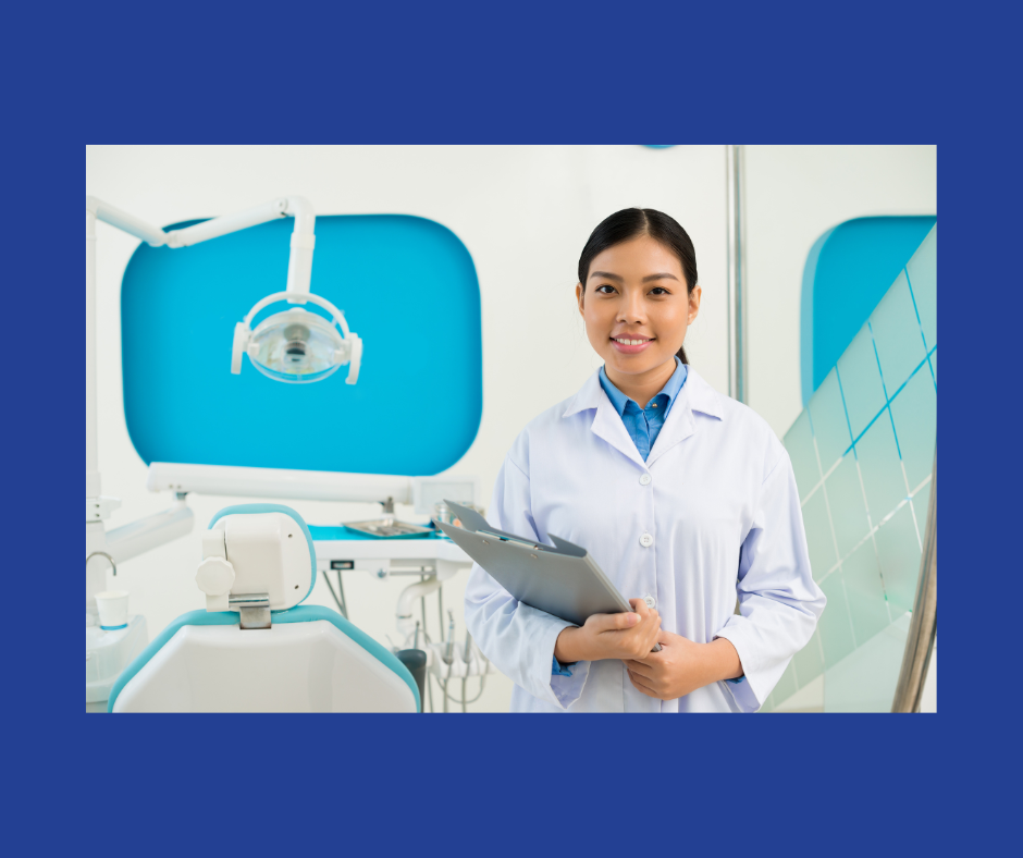 A woman in a white lab coat holding a grey clipboard smiles near a white dental chair and light
