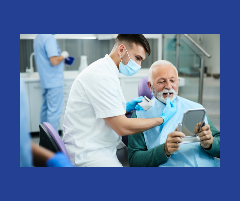 A man in white scrubs and a blue face mask holds up tooth color samples to the mouth of a man in a blue smock who is looking in a mirror