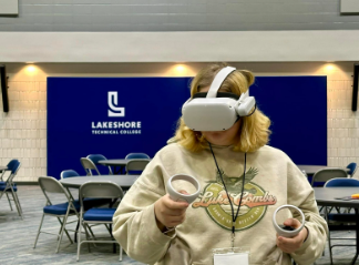 A student with blond hair wears a VR headset in front of a blue sign with white letters that reads, Lakeshore Technical College. 