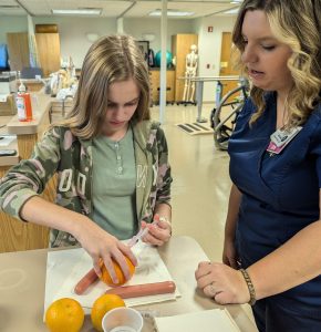 A student learns to give injections by practicing on an orange as an instructor stands by