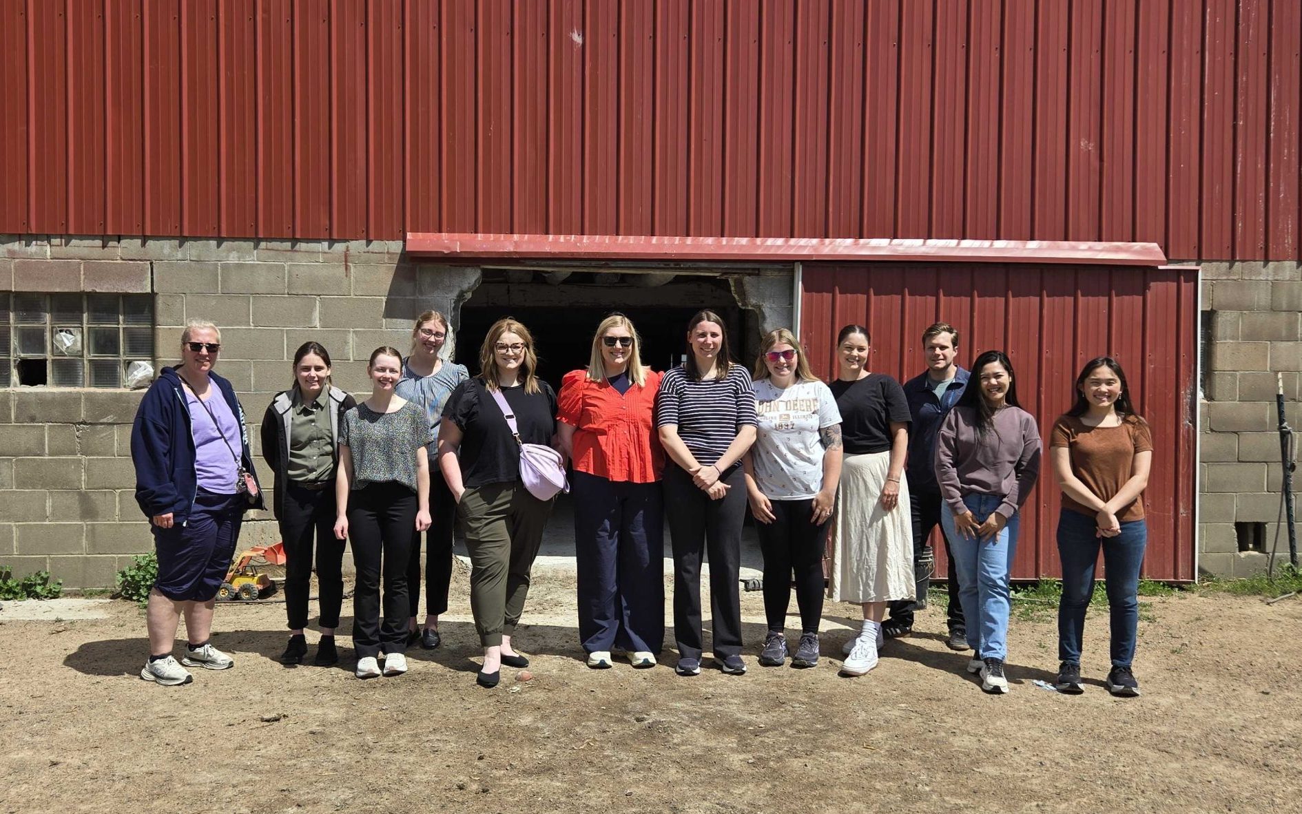 A group of health care students stand in front of a red barn with a grey brick foundation after participating in an AHEC Community Health Immersion