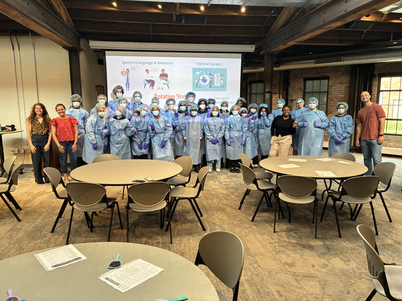 A large group of students in surgical dress pose and smile for the camera in a classroom setting with a projection screen being them. There are tables in front of them with worksheets on top. Two adult instructors stand on either side of the group.